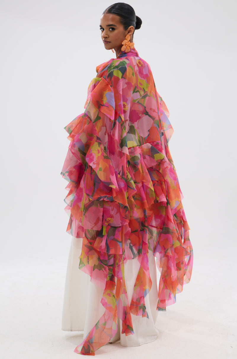 A woman poses in the IN BLOOM FLORAL TOP, a sheer chiffon, ruffled cape with a vibrant pink, orange, and green floral pattern over a white outfit. She styles her hair in a sleek bun and wears bold flower-shaped earrings. 0