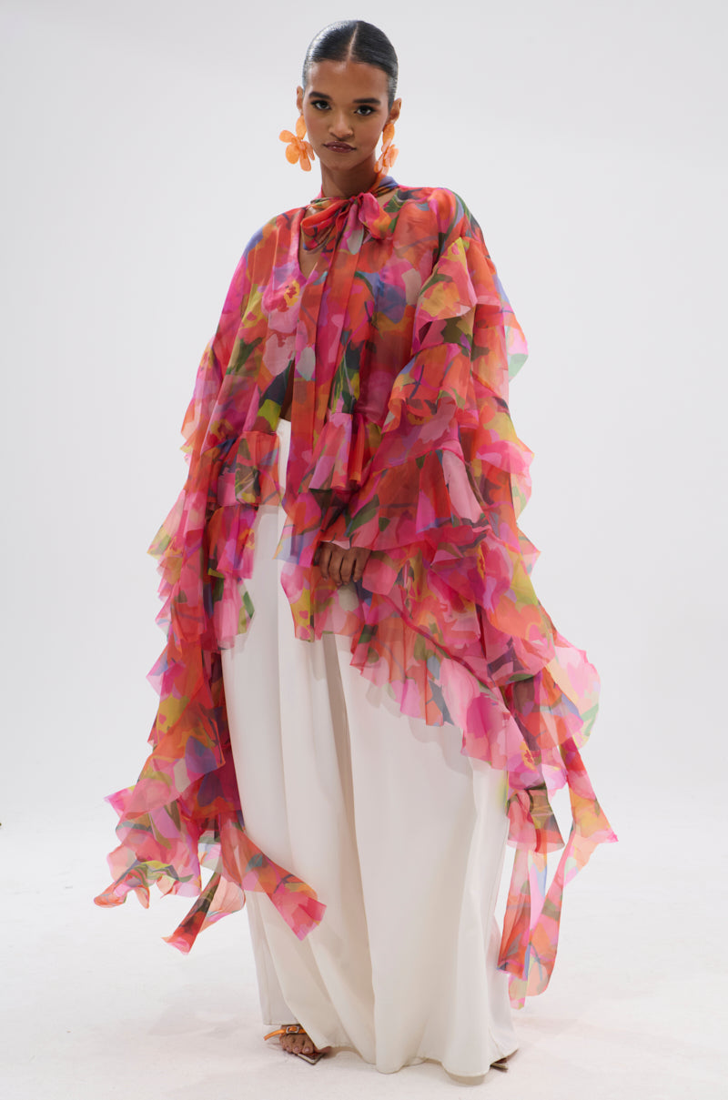 A woman wears the IN BLOOM FLORAL TOP, a colorful sheer ruffled cape with a neck bow, styled as a statement blouse over wide-leg white pants and paired with orange flower earrings against a plain white background. 1