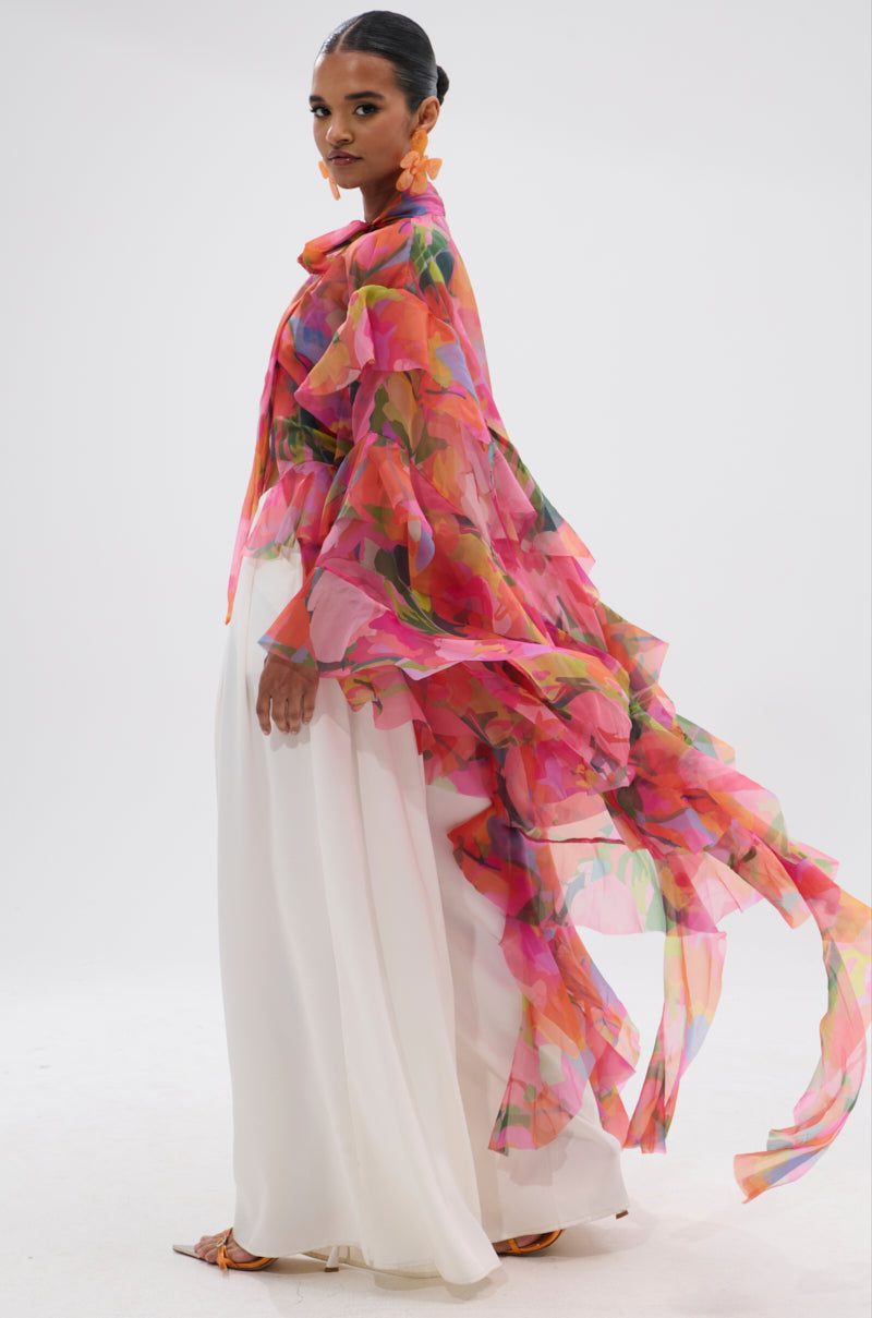 A woman in a sleek bun models the IN BLOOM FLORAL TOP, worn as a sheer, multicolored cape over a long white skirt. The flowing floral fabric billows behind her against a plain white background. 2