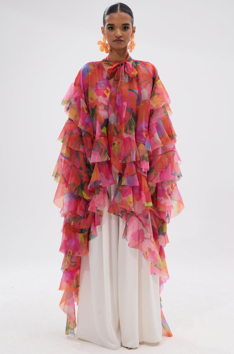 A woman stands against a white background wearing the IN BLOOM FLORAL TOP, a vibrant chiffon ruffle blouse with pink, orange, and yellow floral patterns over wide-legged white pants, finished with large orange flower earrings. 3