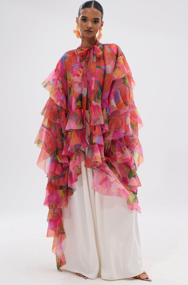 A woman stands against a white background wearing the IN BLOOM FLORAL TOP with vibrant pink, orange, and green ruffles over wide-legged white pants. She completes the look with bold orange earrings and nude heels. 4