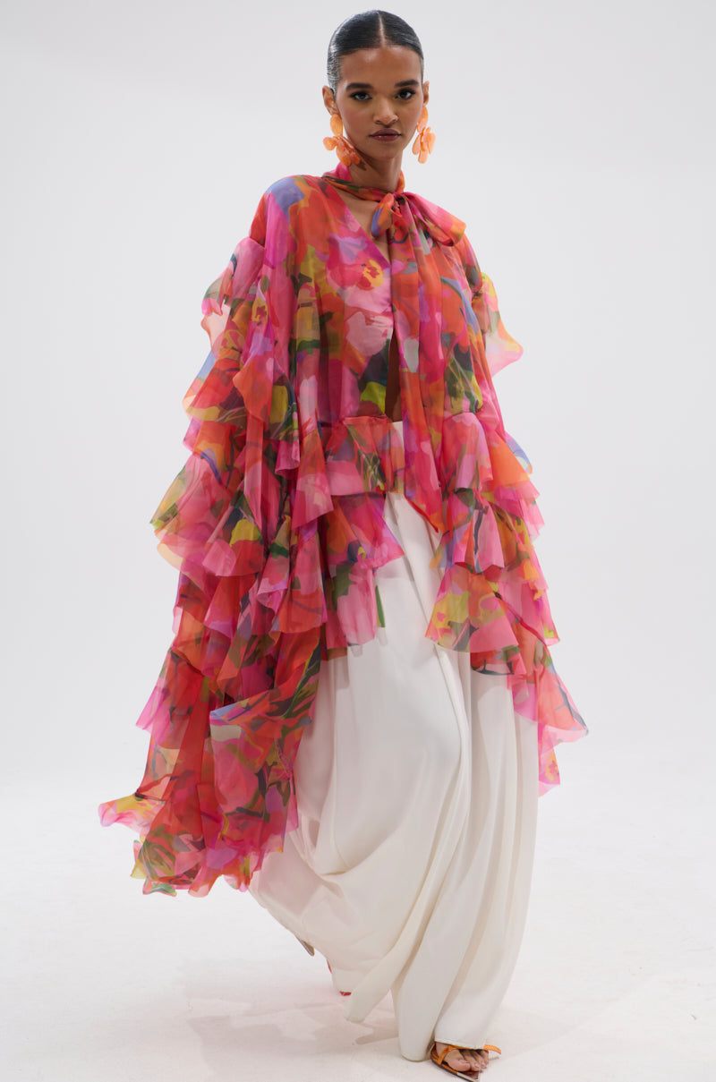 A model wears the IN BLOOM FLORAL TOP—a flowy chiffon ruffle design with pink and orange florals—paired with wide-legged white pants, bold orange earrings, and sandals against a plain white background. 5