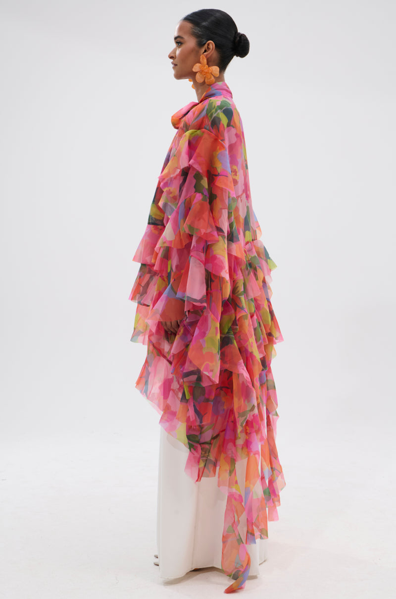 A woman in profile wears the IN BLOOM FLORAL TOP, featuring chiffon ruffles and a colorful floral pattern, paired with white pants. Her hair is styled in a bun and she accessorizes with large orange earrings against a plain background. 6