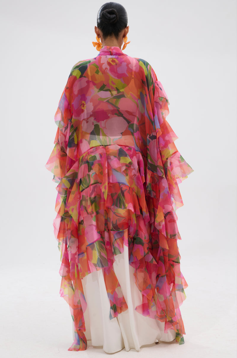A person stands facing away, wearing the IN BLOOM FLORAL TOP—a flowing, sheer blouse with vibrant pink, red, and green florals—over a long white dress against a plain white background. 7