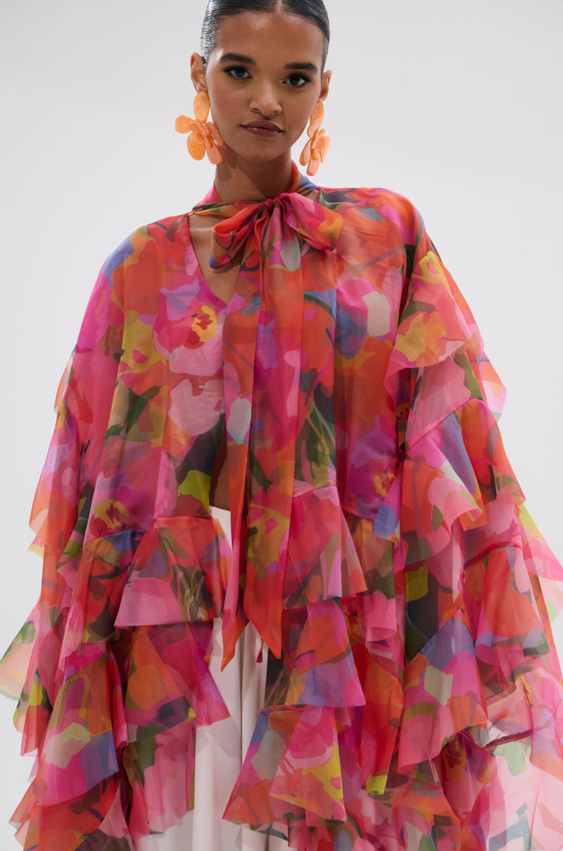 A woman wears the IN BLOOM FLORAL TOP, a sheer chiffon blouse with vibrant floral pattern, ruffle details, and a bow at the neck. She pairs it with orange statement earrings and has her hair in a sleek bun. 8