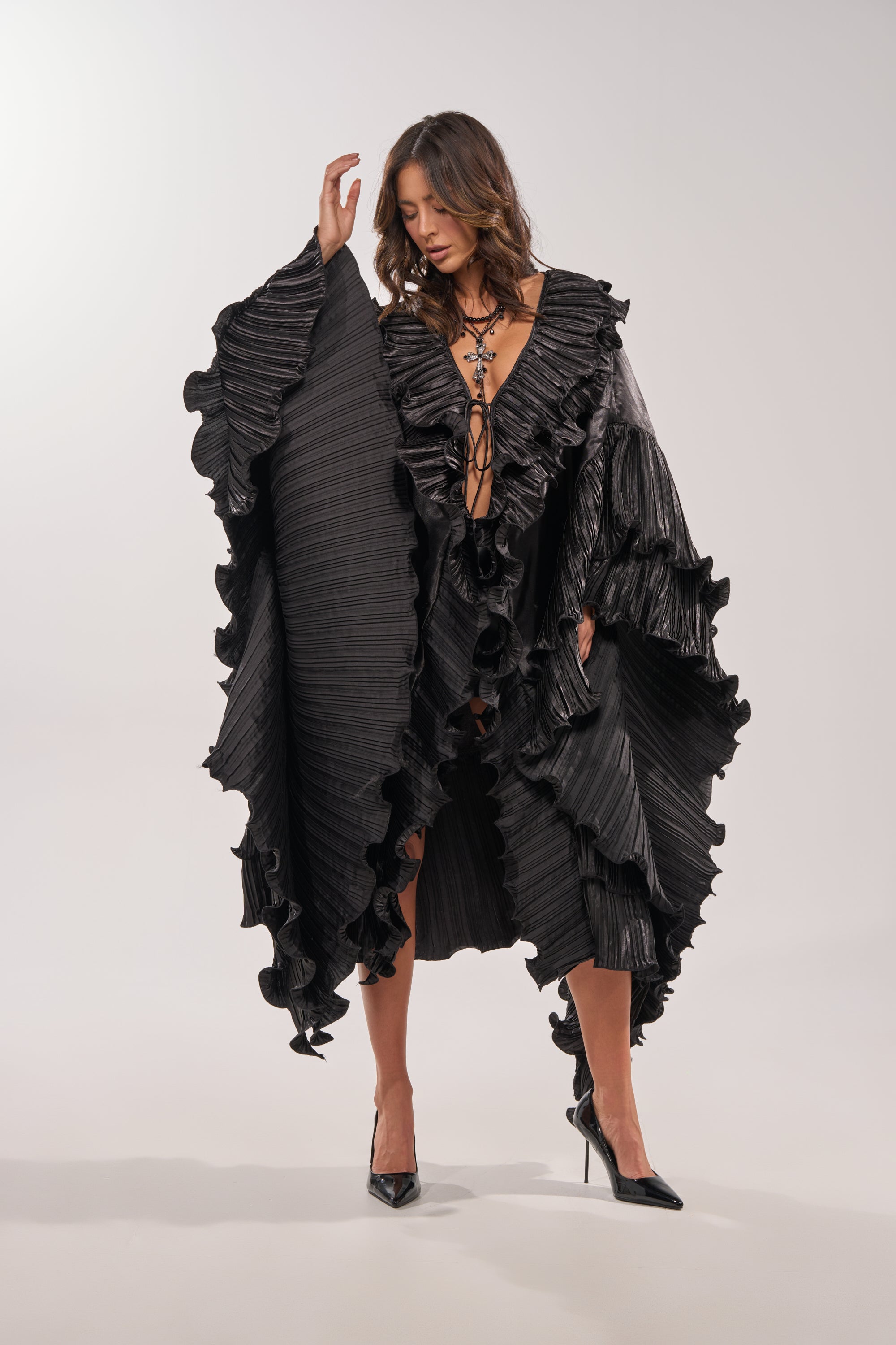A woman models the SHINE ON LONG SLEEVE FRONT TIE TOP styled as a chic black dress with oversized sleeves, deep V-neckline, and black heels against a plain light background. 2