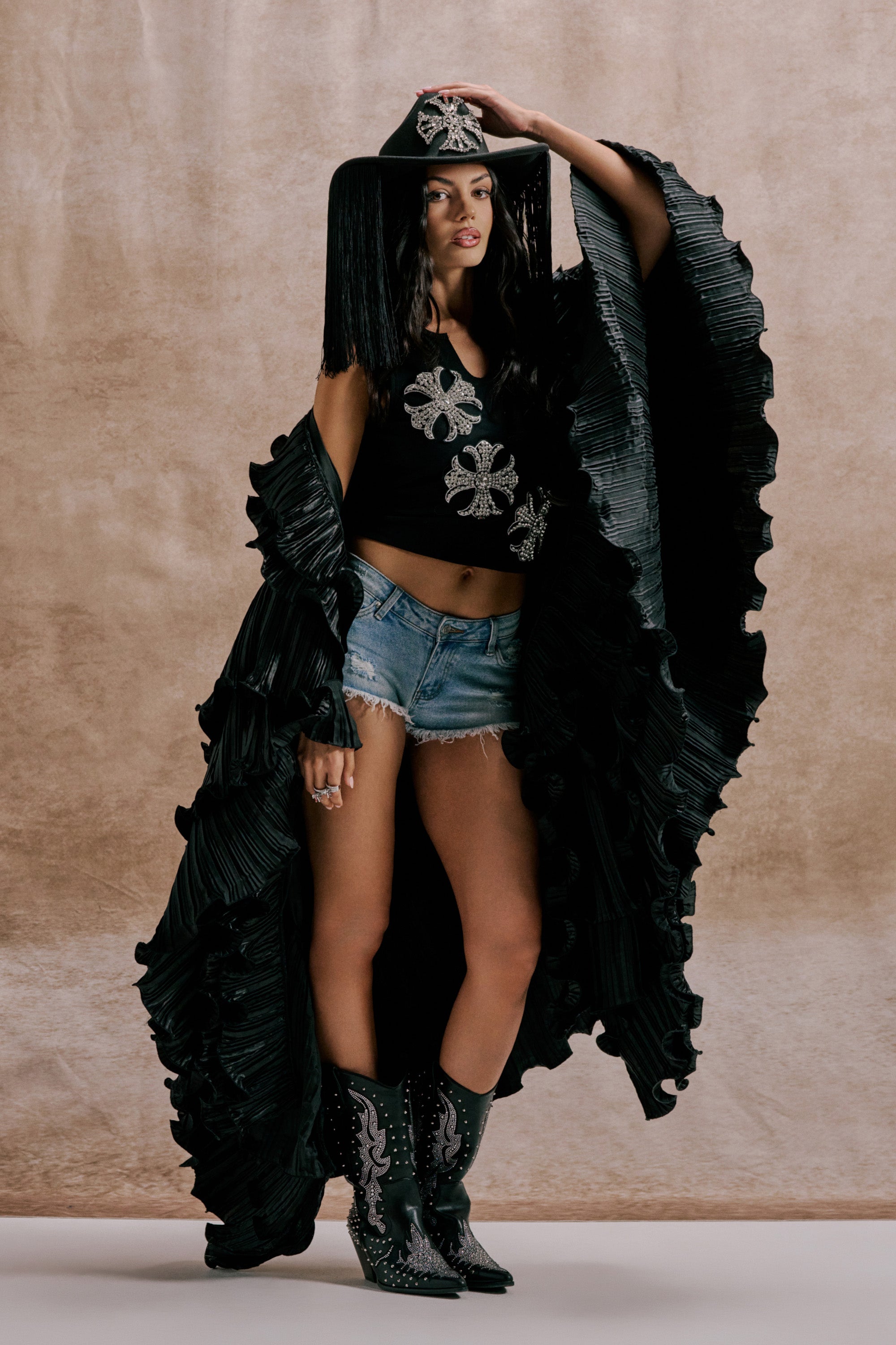 A woman poses in the SHINE ON LONG SLEEVE FRONT TIE TOP with silver embellishments, denim shorts, a black fringe hat, ruffled cape, and black cowboy boots with white embroidery against a neutral textured background. 0