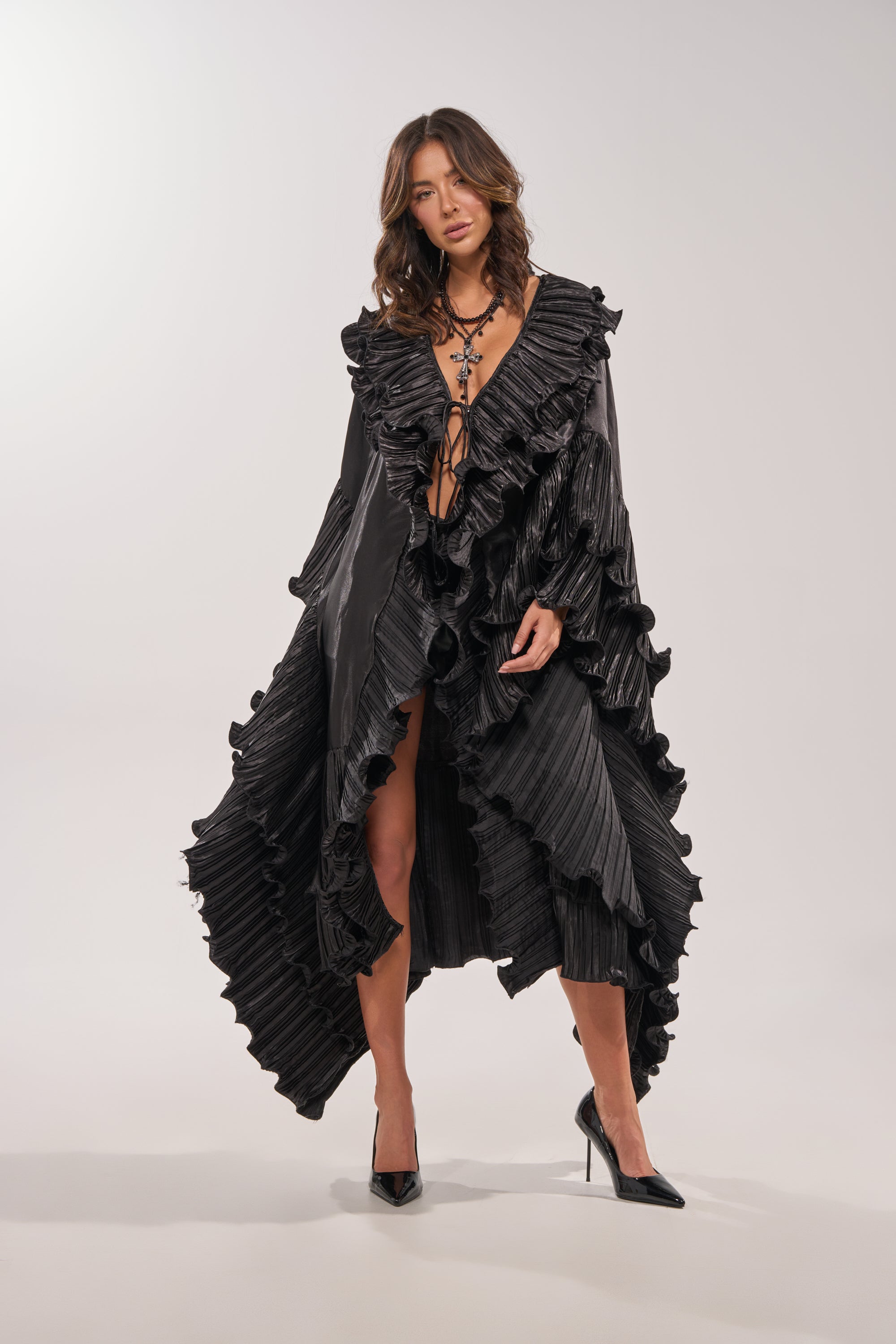 A woman wears the SHINE ON LONG SLEEVE FRONT TIE TOP styled as a black dress, featuring dramatic ruffled edges, a deep V-neckline, and paired with black high heels against a plain light background. 3
