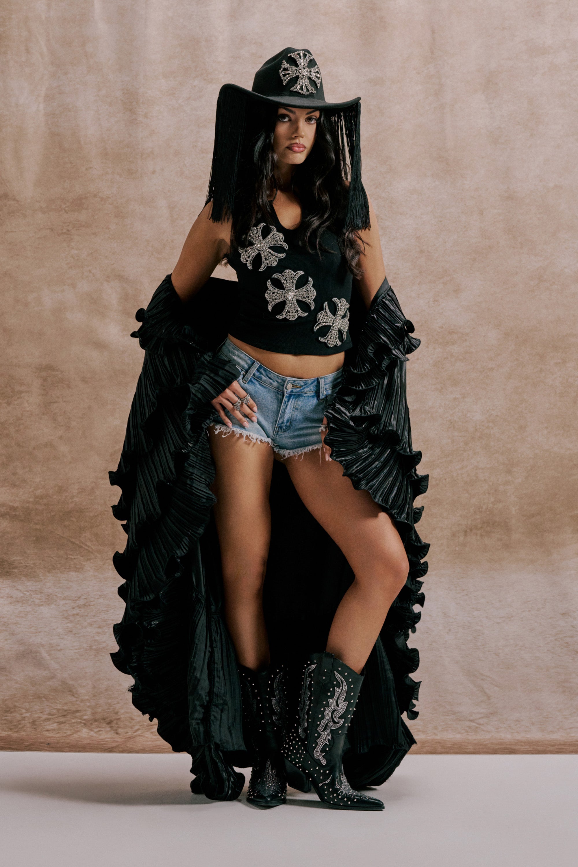A woman stands confidently against a beige backdrop wearing the SHINE ON LONG SLEEVE FRONT TIE TOP, a wide-brimmed black hat with fringe, a ruffled black cape, denim shorts, and black cowboy boots. 1