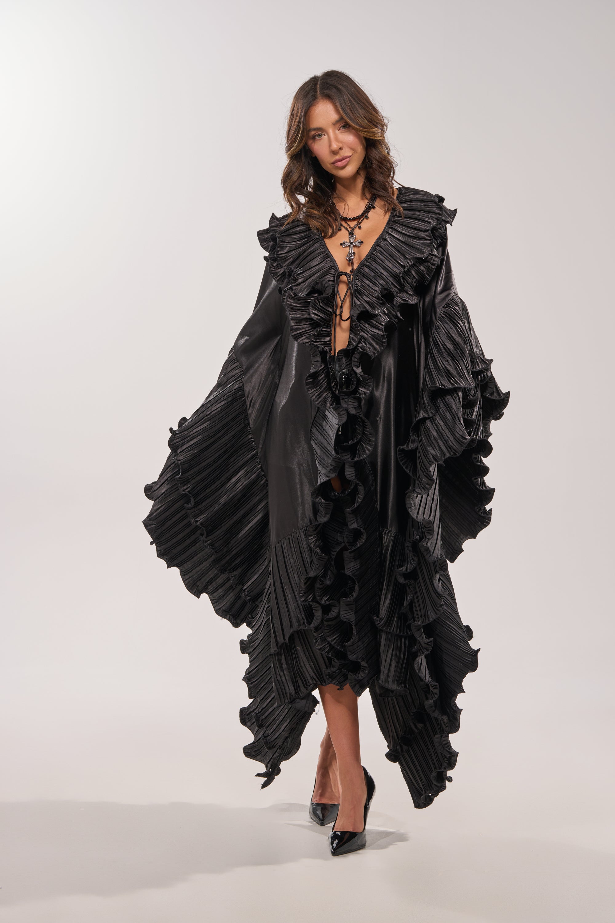 A woman poses in the SHINE ON LONG SLEEVE FRONT TIE TOP, a dramatic black blouse with pleated ruffle edges, a deep V-neckline, and oversized sleeves. She pairs it with black heels and stands against a plain background, her wavy brown hair down. 4