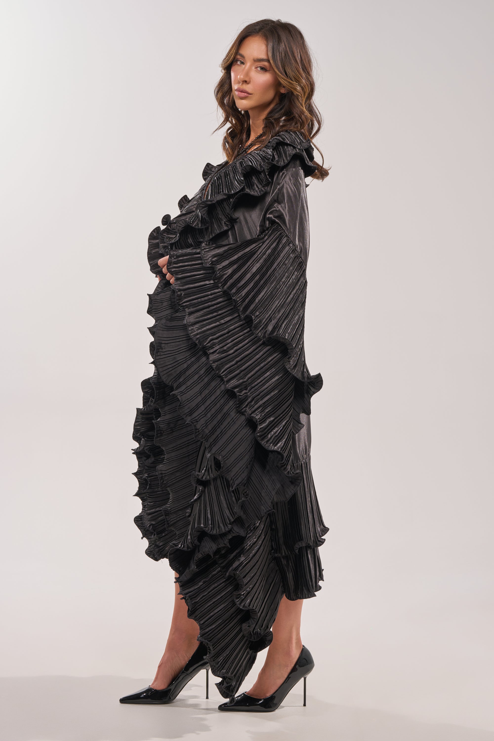 A woman with wavy brown hair and black heels poses in profile against a light background, wearing the SHINE ON LONG SLEEVE FRONT TIE TOP styled as a dramatic cascading black dress with pleated ruffles. 6