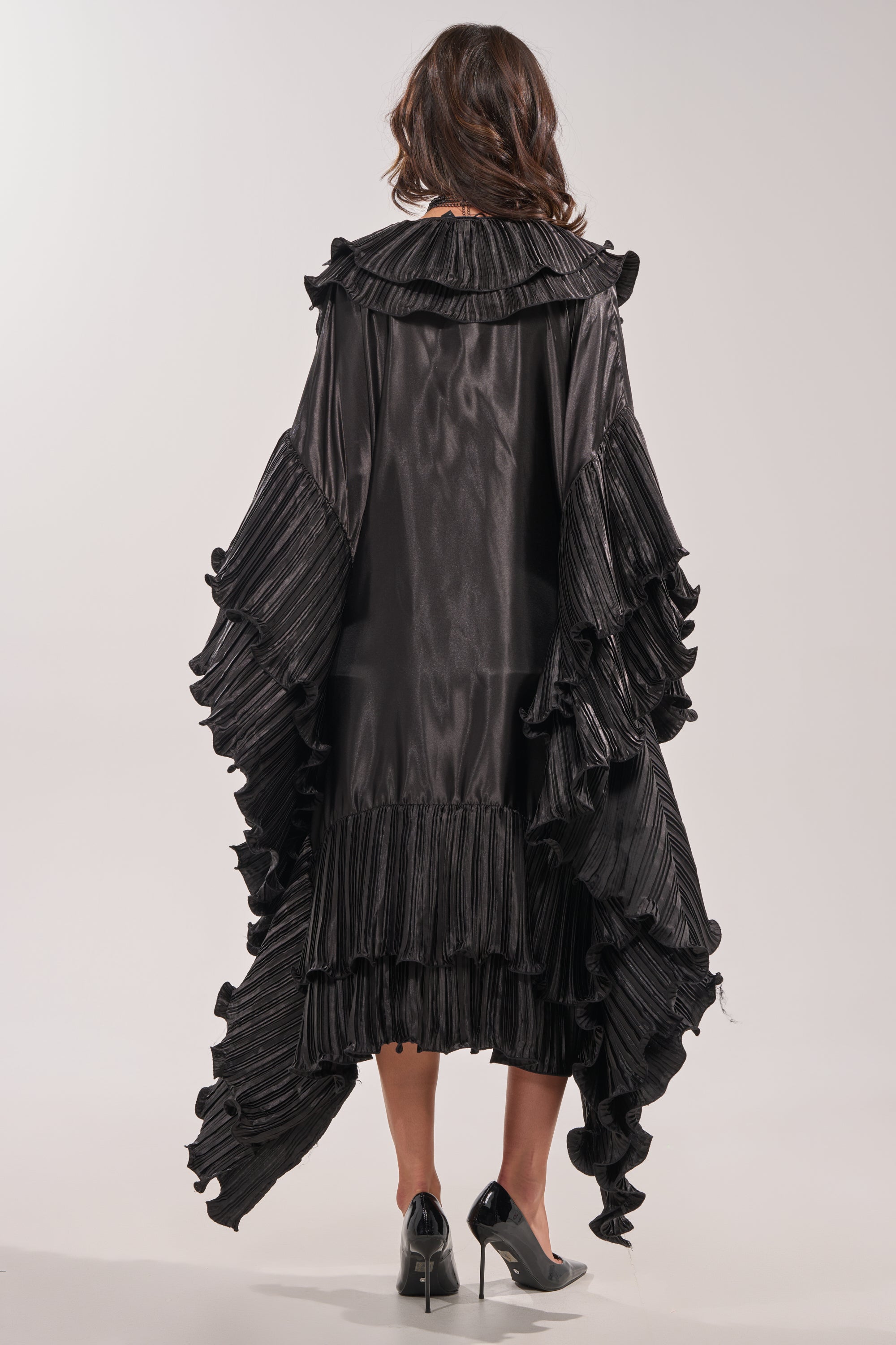 A woman with wavy brown hair and black heels faces away, wearing the SHINE ON LONG SLEEVE FRONT TIE TOP—a striking black blouse featuring ruffled, pleated layers and a high-low hemline. 8