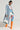 A person is dressed in a long, light blue coat over the vibrant FEELIN MYSELF PRINTED JUMPSUIT, which features an abstract graphic print and a bodycon fit. They pair this ensemble with striking bright orange knee-high boots and carry a white handbag. Completing the look with blue-tinted glasses, they stand confidently in a bright, minimalist setting.