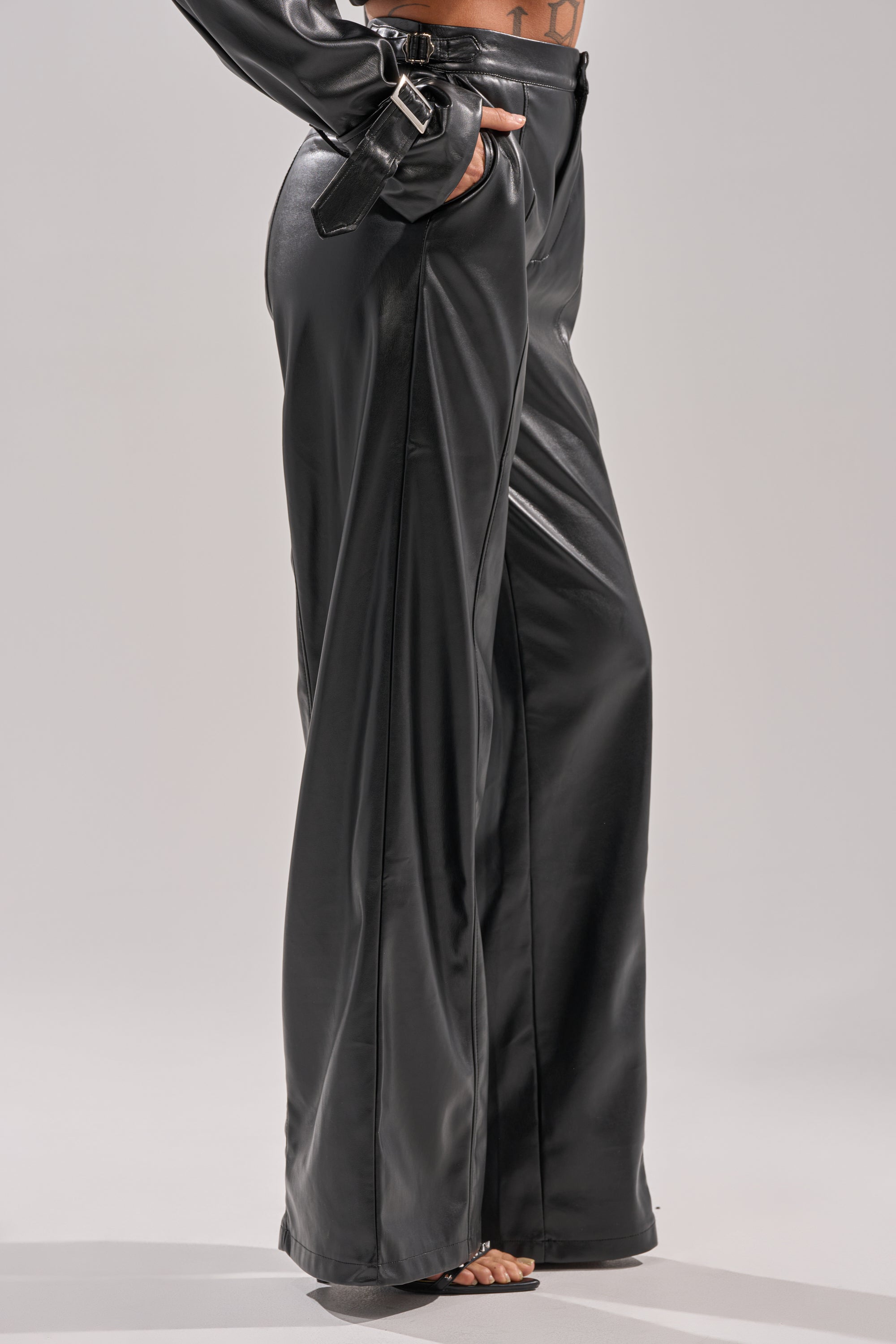 A person models the MONTE CARLO WIDE LEG PANT IN BLACK, featuring a high-rise fit and a matching faux leather belt. Only the lower body in these chic black faux leather wide leg pants is shown against a plain light background.