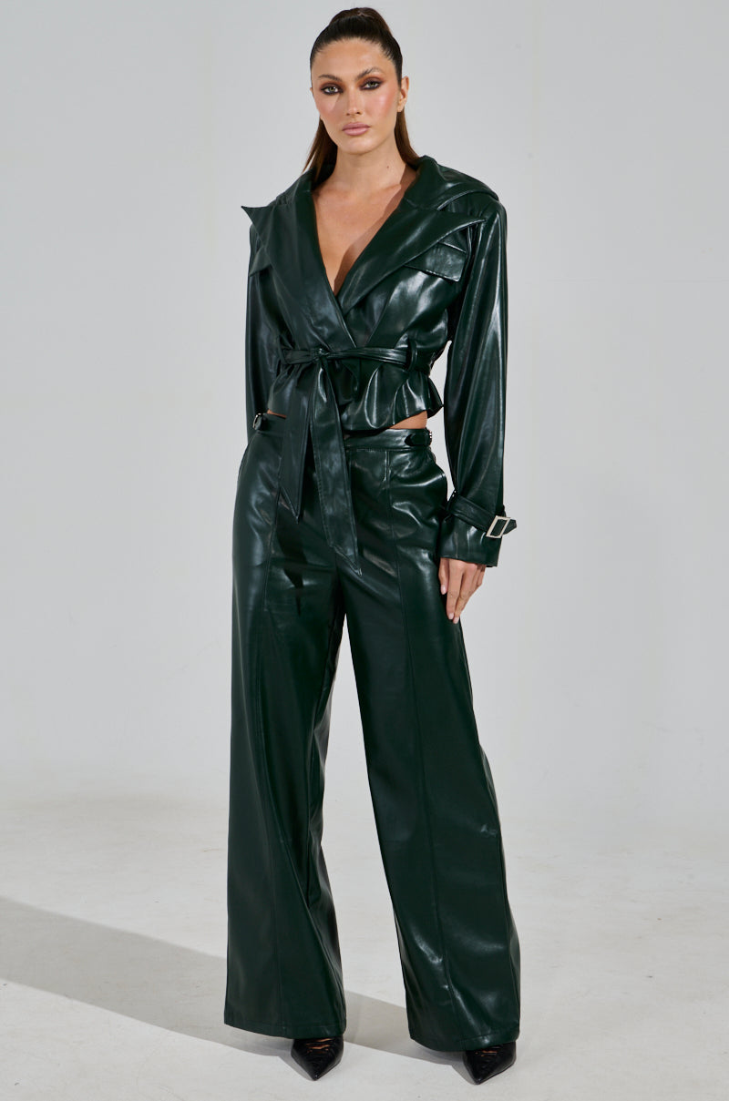 A person stands confidently in an evergreen-hued, belted MONTE CARLO HOODED MOTO faux leather jacket and wide-leg pants. Their long hair is pulled back, they wear black pointed shoes, and the background is plain and light-colored.