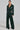A person stands confidently in an evergreen-hued, belted MONTE CARLO HOODED MOTO faux leather jacket and wide-leg pants. Their long hair is pulled back, they wear black pointed shoes, and the background is plain and light-colored.