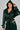 A person poses against a plain background wearing the MONTE CARLO HOODED MOTO in dark green faux leather, featuring a cropped design, matching pants, a waist sash, and button-detailed sleeves.