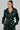 A person is wearing the MONTE CARLO HOODED MOTO in dark green faux leather with a belt, along with matching pants. They have long, dark hair in a high ponytail and are standing with hands in pockets against a plain background.