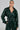 A person confidently wears the MONTE CARLO HOODED MOTO in dark green faux leather with matching pants. The cropped jacket, featuring a detachable belt at the waist, complements their long, sleek hair against a plain, light-colored background.