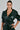 Against a plain background, a person with long, dark hair pulled back confidently models the MONTE CARLO HOODED MOTO—an evergreen faux leather jacket featuring a belted waist and deep neckline.
