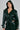 Wearing the MONTE CARLO HOODED MOTO, a chic dark green faux leather jacket with a belted waist and layered lapels, paired with matching pants against a light gray backdrop.