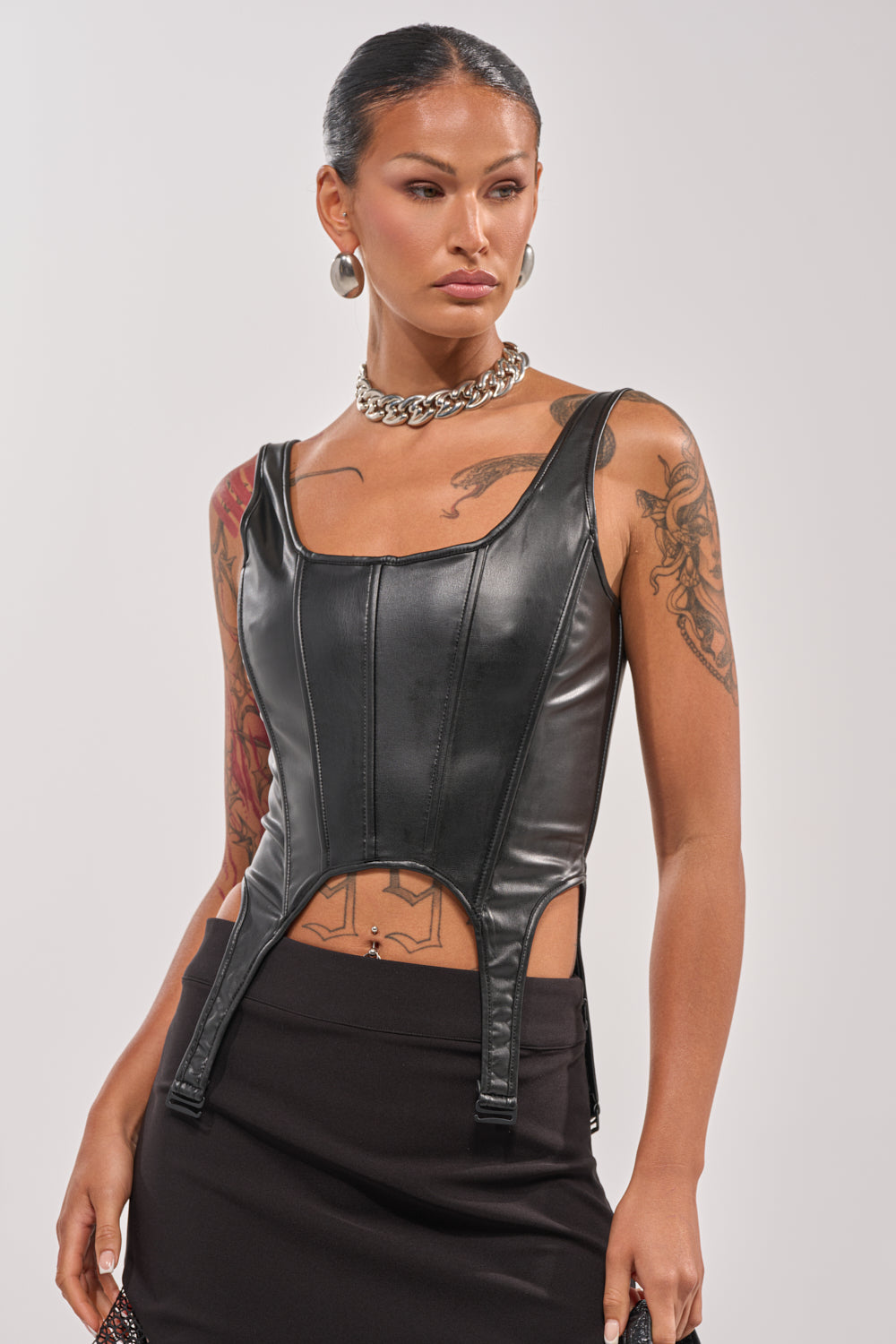 A person with tattoos and slicked-back hair wears the LOCKED AND LOADED FAUX LEATHER CORSET, featuring subtle garter strap details, styled with a black skirt, silver chain necklace, and hoop earrings against a light background. 0
