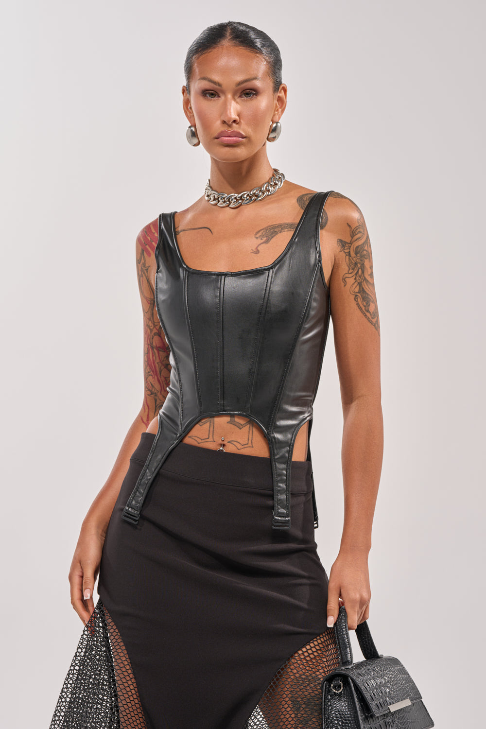 A person in the LOCKED AND LOADED FAUX LEATHER CORSET, black mesh-panel skirt, chunky chain necklace, hoop earrings, and holding a black textured handbag. Arm and chest tattoos add edge to this lingerie-inspired look. 1