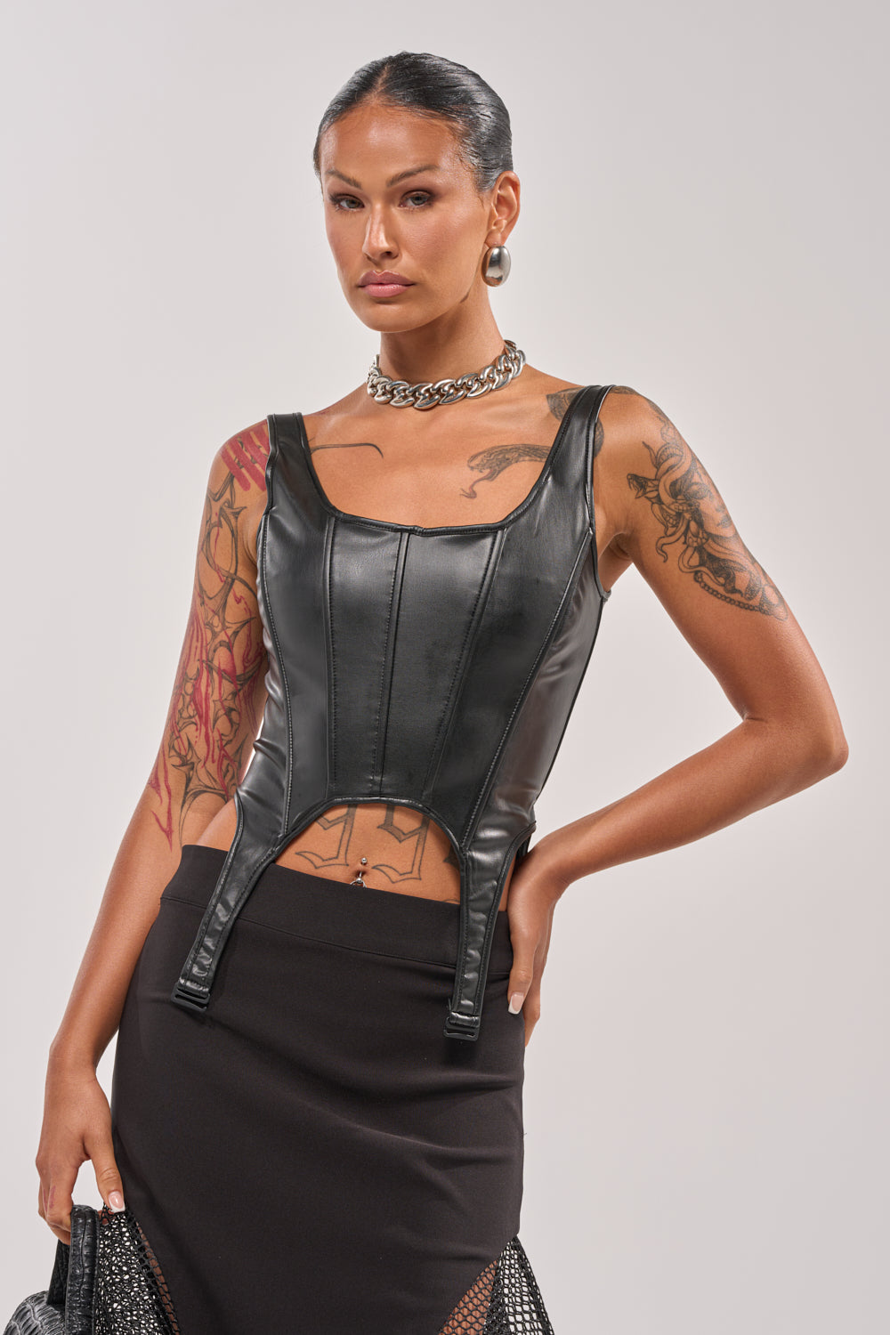 A person with slicked-back hair poses confidently in the LOCKED AND LOADED FAUX LEATHER CORSET and a black skirt, accessorized with a chunky chain necklace and hoop earrings, showing off arm tattoos against a plain background. 2