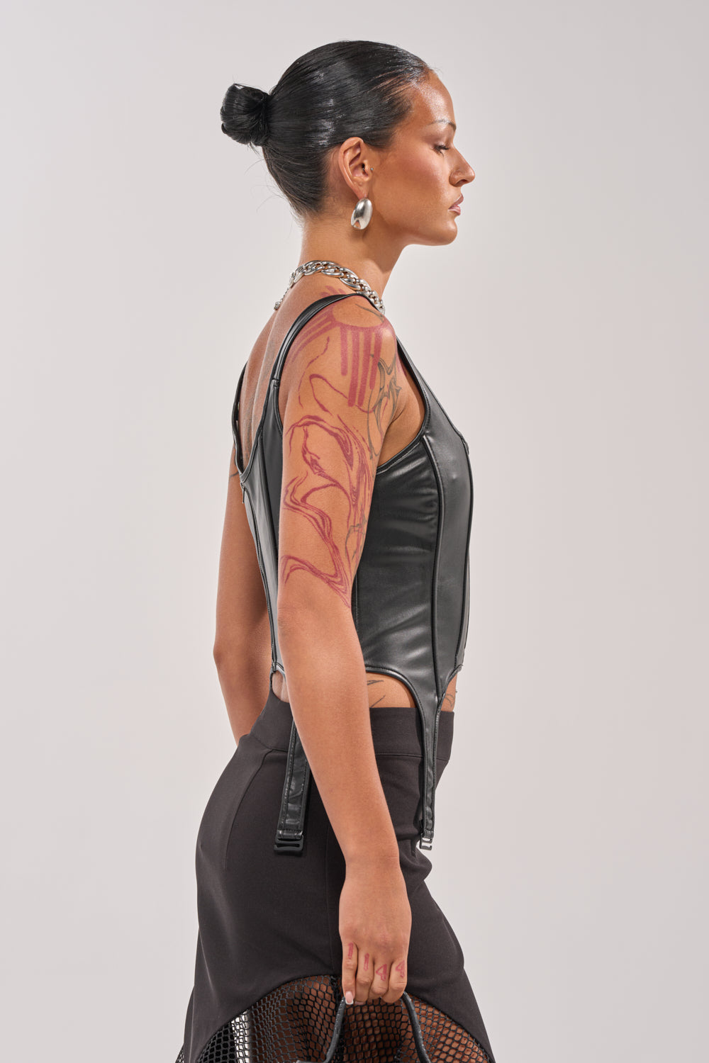 A woman with slicked-back hair in a bun and a red abstract upper arm tattoo stands in profile, styled with the LOCKED AND LOADED FAUX LEATHER CORSET, black skirt, chunky silver jewelry, and a black bag. 3
