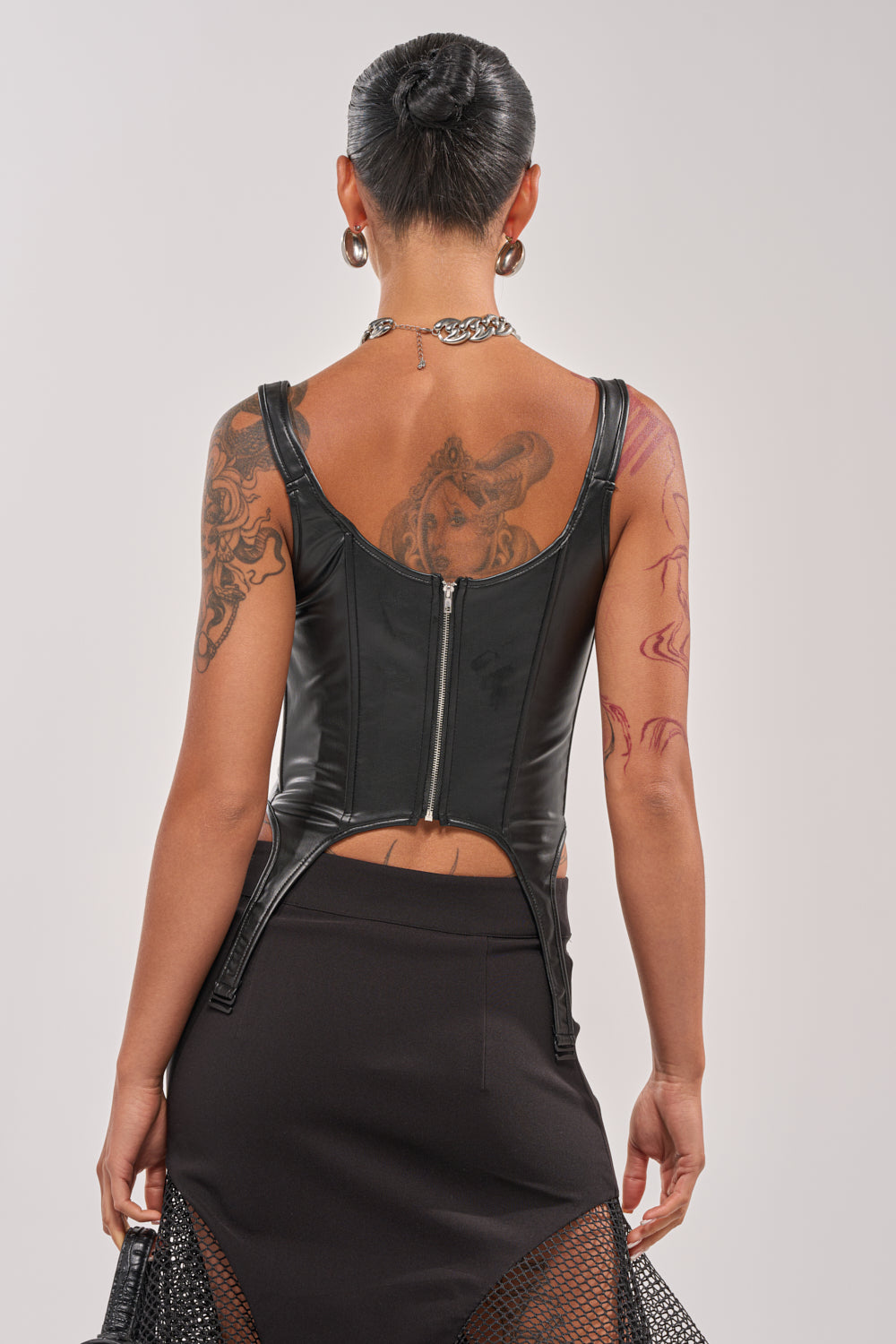 A person with short dark hair, visible tattoos, and hoop earrings is shown from the back wearing the LOCKED AND LOADED FAUX LEATHER CORSET in black, paired with a black skirt featuring mesh panels. 4