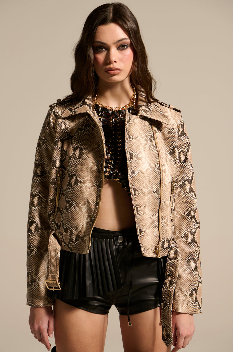 Wearing the REBOOT SNAKE MOTO jacket over a black top and black pleated leather shorts, someone with long, wavy hair stands calmly against a neutral background, facing the camera.