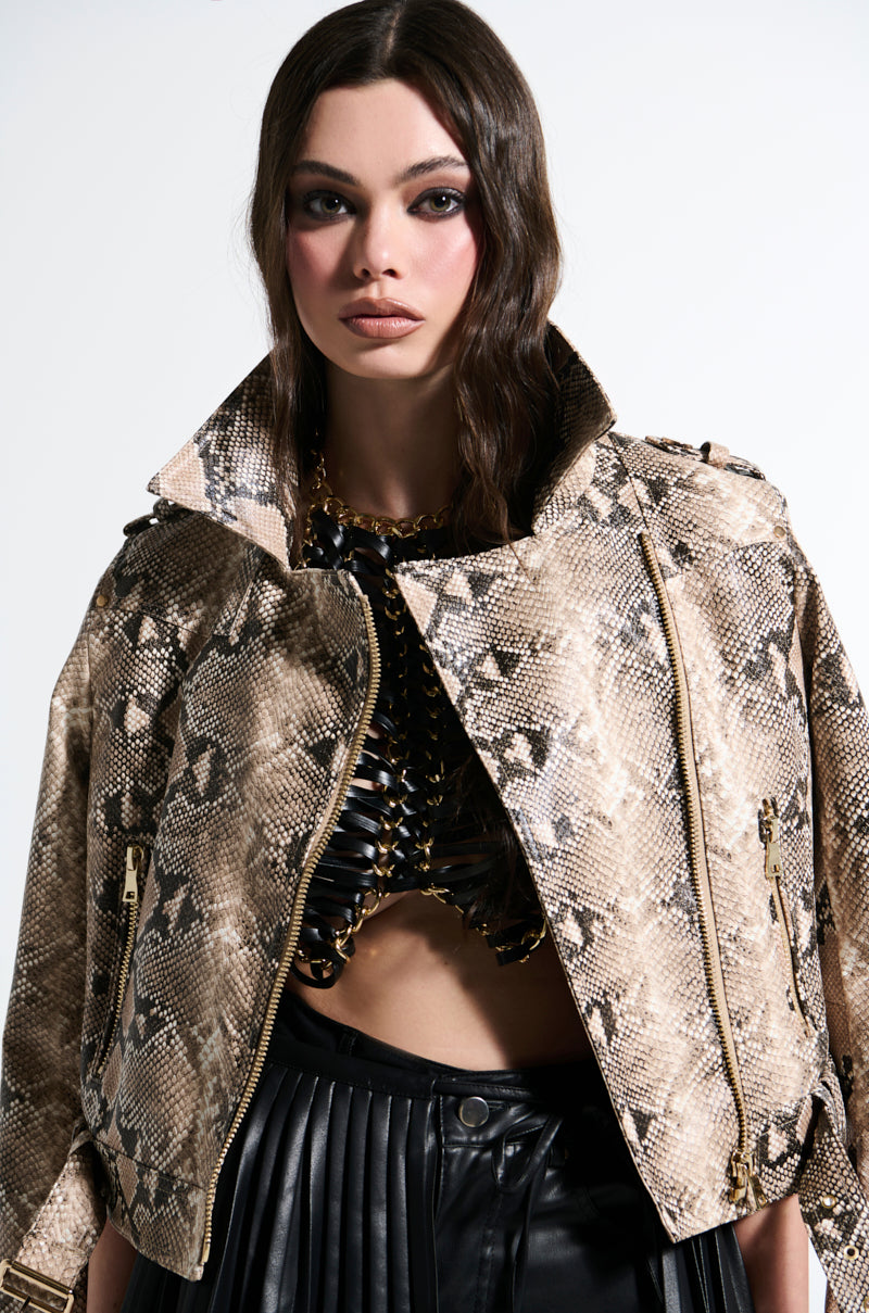 A woman with long dark hair wears the REBOOT SNAKE MOTO jacket over a black textured top and dark pleated pants, exuding sophistication. Against a light background, her neutral expression highlights the faux leather jacket's embossed snakeskin print.