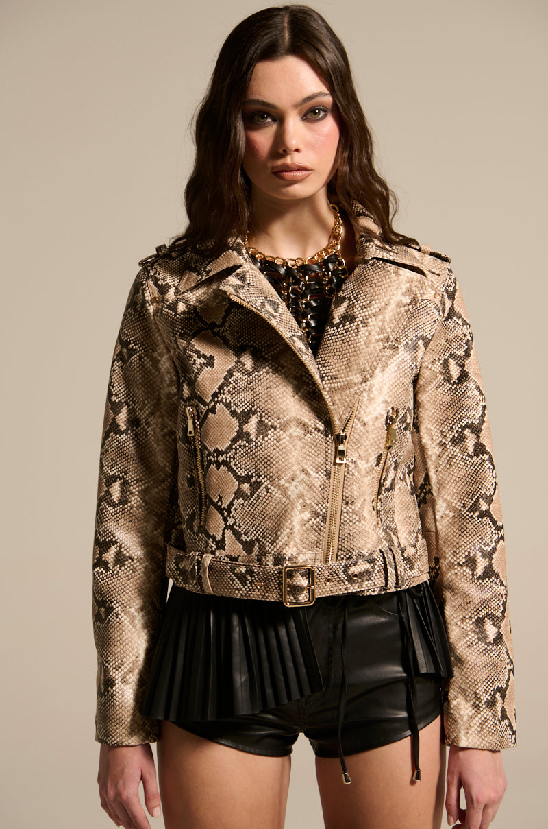 A woman wears the REBOOT SNAKE MOTO jacket, featuring zippers and a belt, styled with black shorts and pleated hem. Her long dark wavy hair perfectly complements her chunky necklace against the plain background.