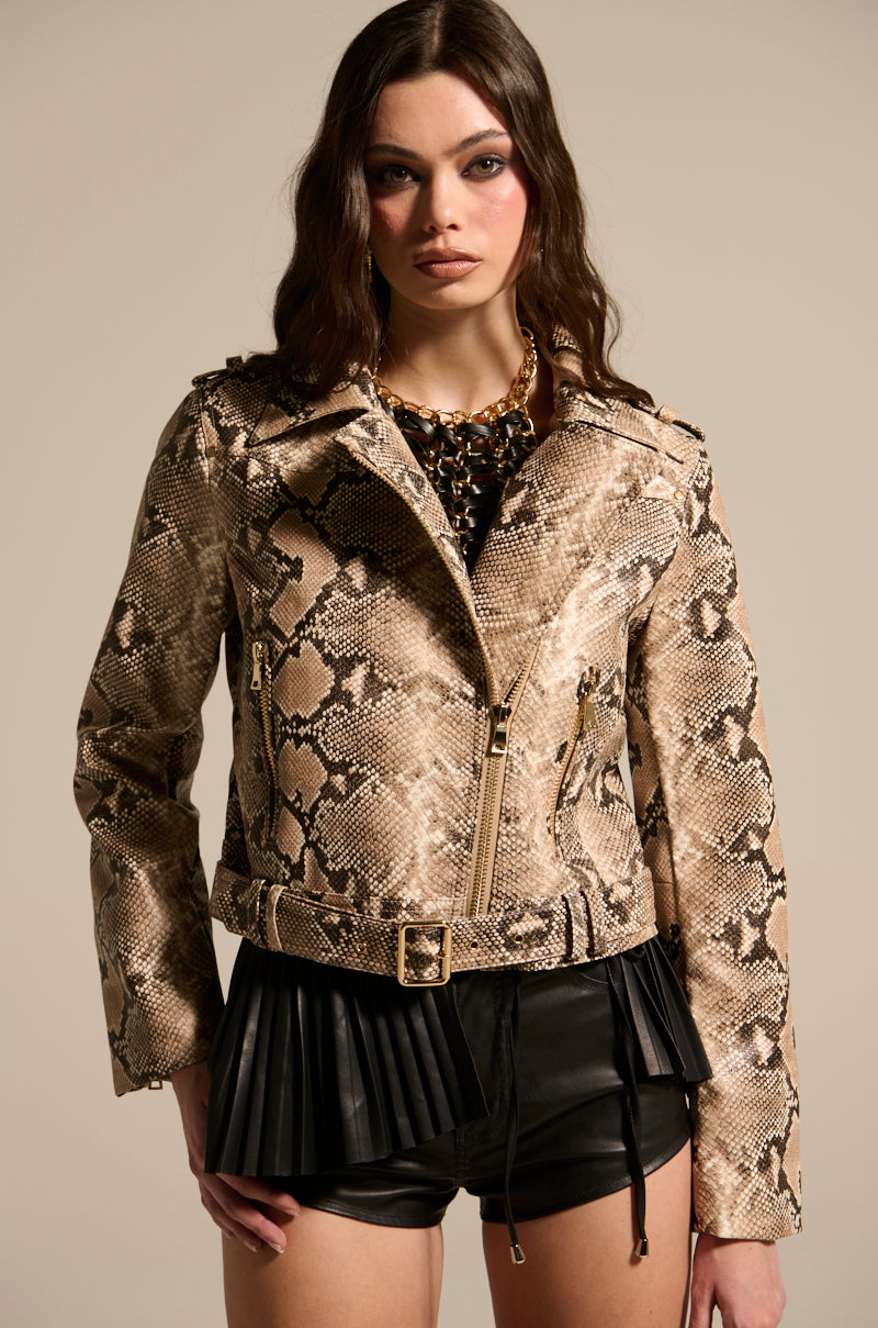 Wearing the REBOOT SNAKE MOTO jacket with embossed snakeskin print and black leather shorts, a person stands confidently against a neutral background. The outfit showcases pleated leather details and gold zipper accents, complementing their long, wavy hair.