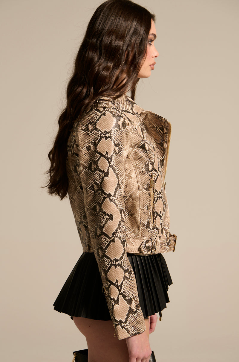 A person with long brown hair is shown in profile, wearing a REBOOT SNAKE MOTO jacket and a black pleated mini skirt against a plain beige background.