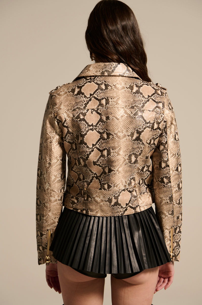 A person with long hair wears the REBOOT SNAKE MOTO, an embossed snakeskin-print faux leather jacket, paired with a black leather pleated skirt. They stand with their back to the camera against a plain beige background.