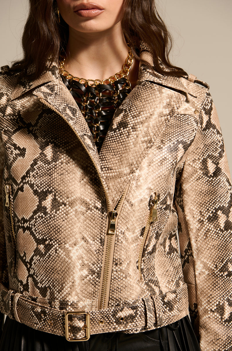 A person is flaunting the REBOOT SNAKE MOTO, a beige and black embossed snakeskin print jacket with gold zippers and a belt. It's layered over a sleek black top and accessorized with a chunky gold chain necklace. Only the torso and part of the face are visible.