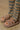 A close-up shows a person wearing the AZALEA WANG HONOR TAN SANDALS from the loungewear collection, paired with striped green and brown pants. The open almond toe design highlights their light-painted toenails as they stand on a concrete surface.