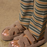 A close-up shows a person wearing the AZALEA WANG HONOR TAN SANDALS from the loungewear collection, paired with striped green and brown pants. The open almond toe design highlights their light-painted toenails as they stand on a concrete surface.