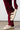 Dressed in a burgundy knit outfit, a person adjusts the AZALEA WANG HONOR WHITE SANDAL, made of fluffy cream-colored sherpa material, on their foot. This cozy open-toe sandal features three straps and is placed on a neutral floor against a plain white background.