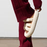 Dressed in a burgundy knit outfit, a person adjusts the AZALEA WANG HONOR WHITE SANDAL, made of fluffy cream-colored sherpa material, on their foot. This cozy open-toe sandal features three straps and is placed on a neutral floor against a plain white background.