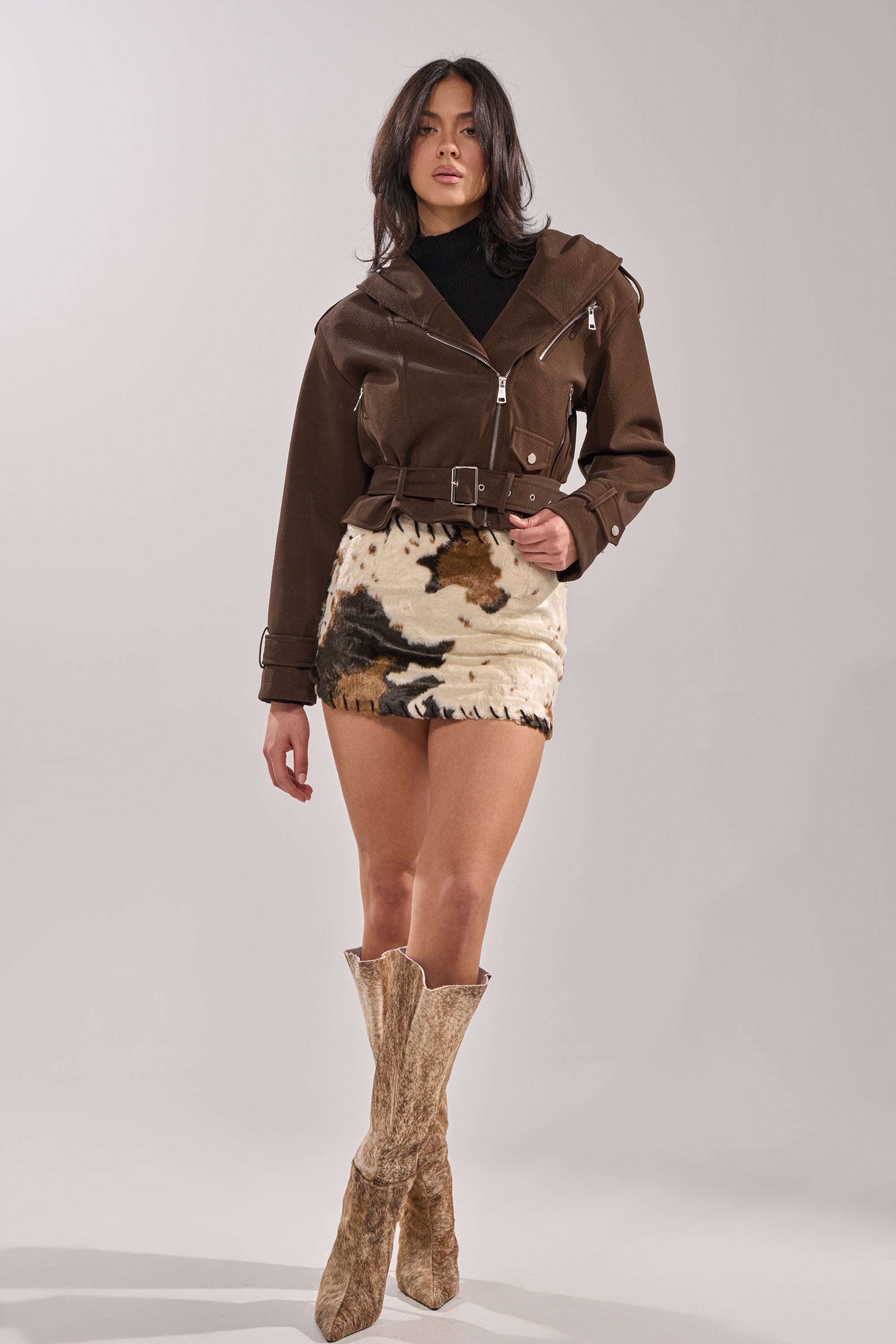 A woman stands in a studio wearing the KIRA SUEDE EFFECT HOODED MOTO jacket, a black turtleneck, cow-print mini skirt, and tan knee-high boots. She faces forward with one arm relaxed and the other slightly bent. 0