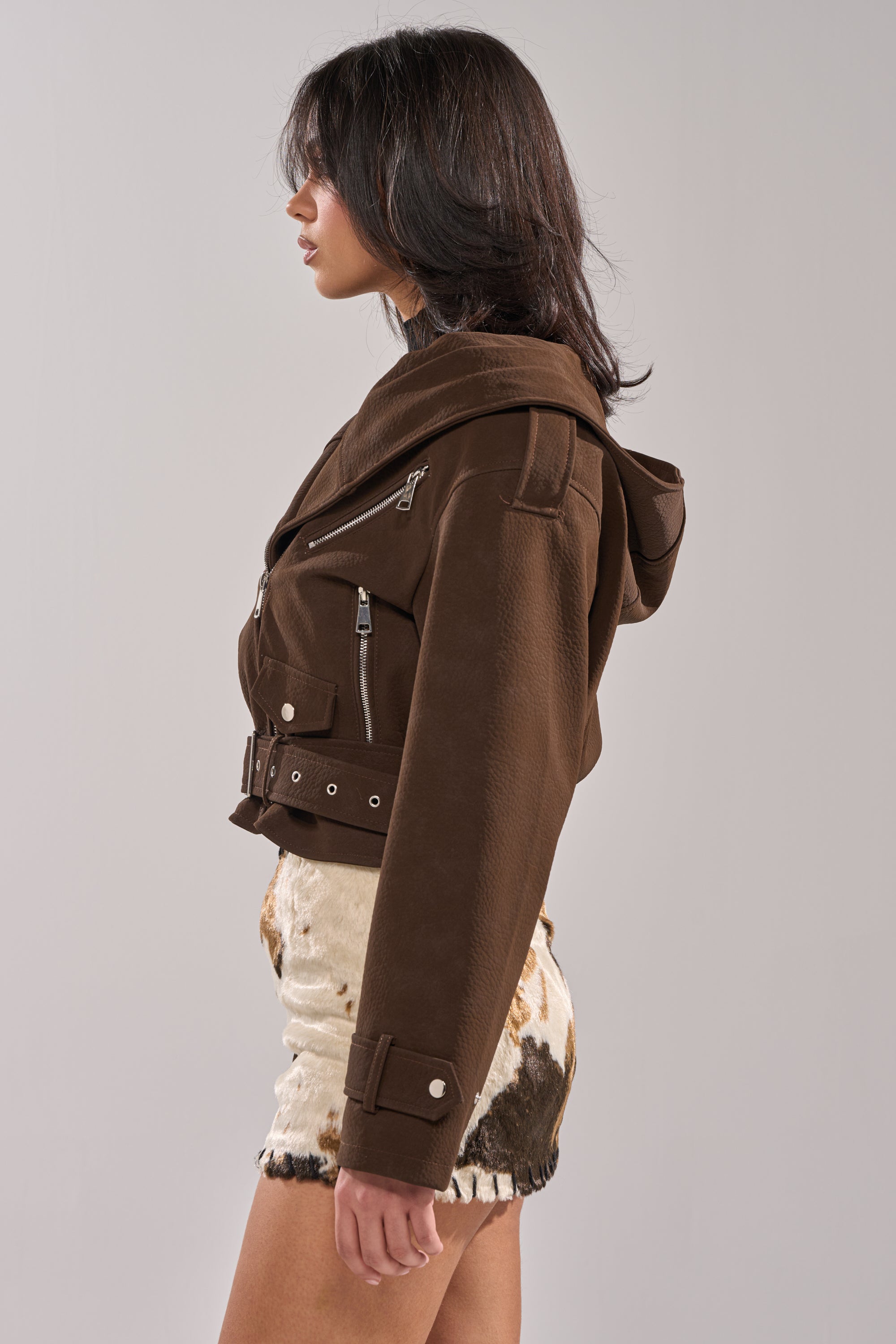 A woman in profile wears the KIRA SUEDE EFFECT HOODED MOTO, a brown faux suede moto jacket with silver zippers, and light cow-print shorts against a plain background. 2