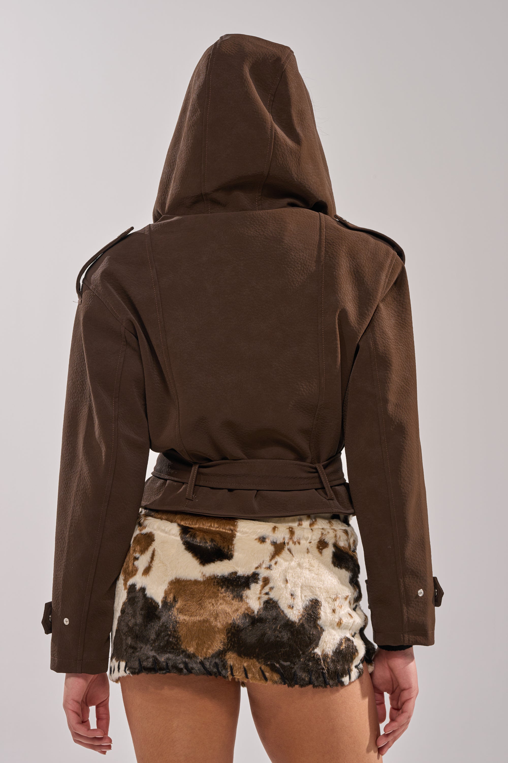 Someone stands with their back to the camera, wearing the KIRA SUEDE EFFECT HOODED MOTO jacket and a short, multicolored furry skirt with a cowhide pattern, set against a plain background. 3