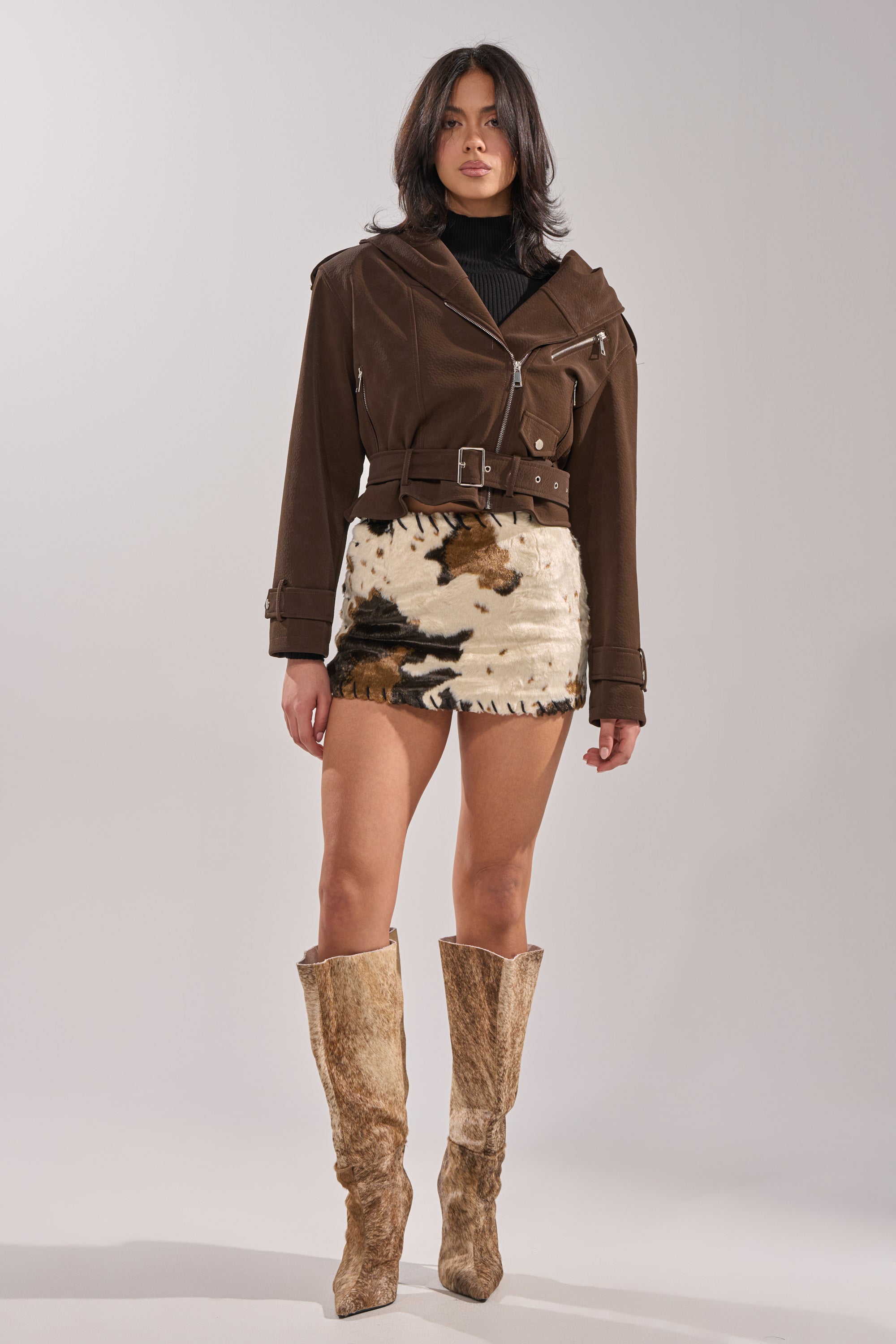 A woman faces forward against a plain light background, wearing the KIRA SUEDE EFFECT HOODED MOTO jacket over a black turtleneck, a cow-print mini skirt, and tall brown textured boots. Her expression is neutral. 4