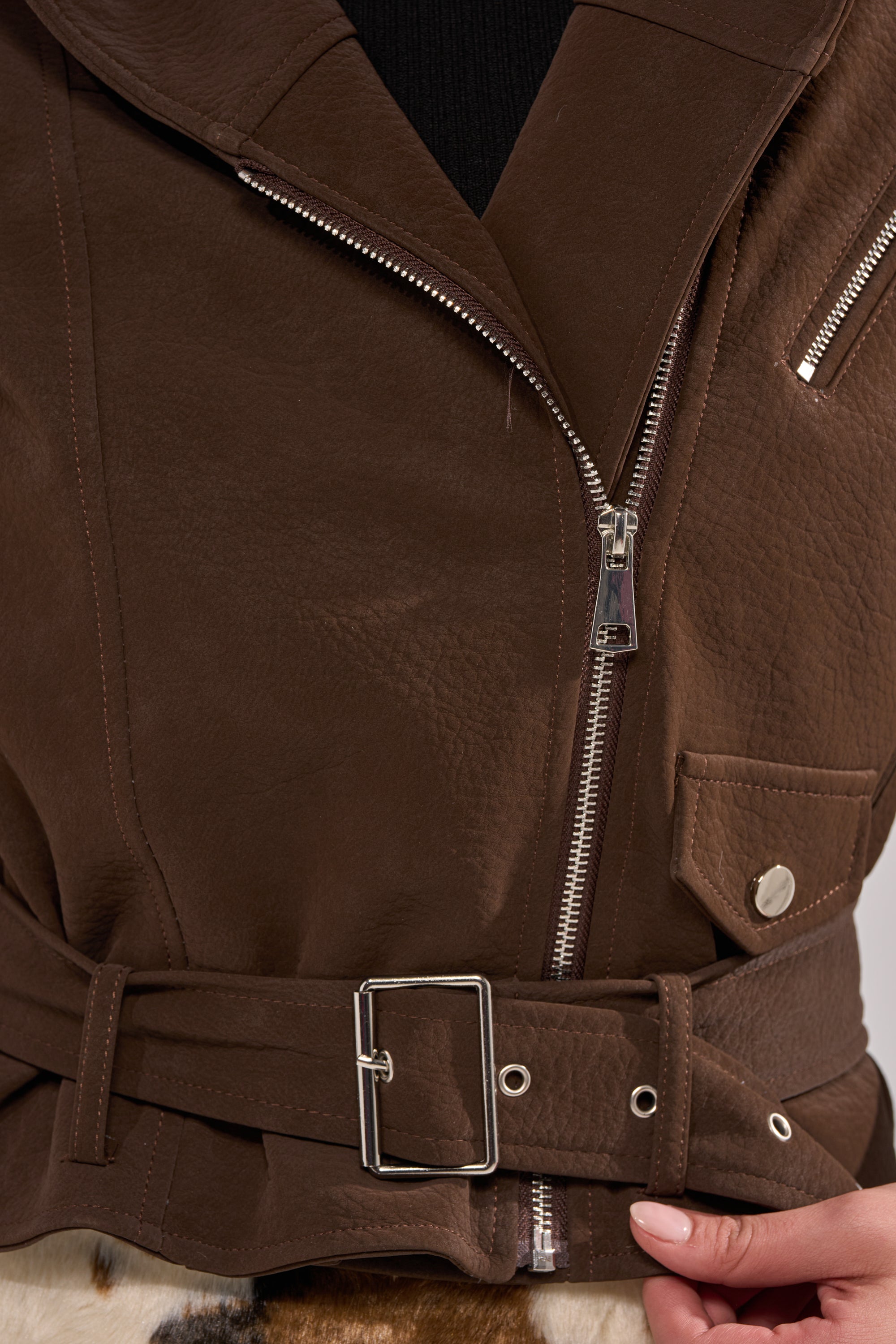 A close-up of the KIRA SUEDE EFFECT HOODED MOTO, a brown faux leather jacket with silver zippers, buckle belt, and a textured finish, worn over a black top. A hand grips the belt for an edgy streetwear vibe. 5
