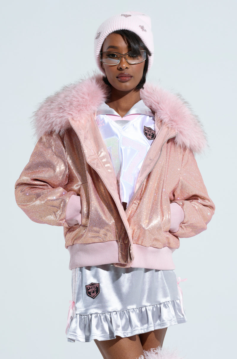 A person is wearing the "Eyes On Me Rhinestone Puffer" jacket with a detachable faux fur collar, paired with a white top and a silver skirt featuring pink details. Their pink beanie, decorated with heart designs, and glasses add charm against the plain background.