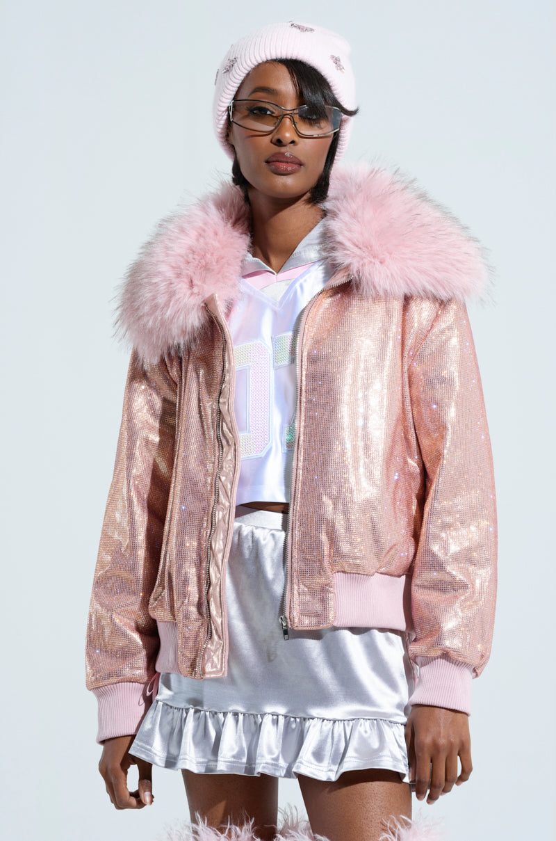 A person wearing the "Eyes On Me Rhinestone Puffer" jacket with a detachable faux fur collar, a white shirt, a silver metallic skirt, a pink beanie, and sunglasses. This ensemble expertly combines various textures and soft tones to achieve an elegant and contemporary appearance.