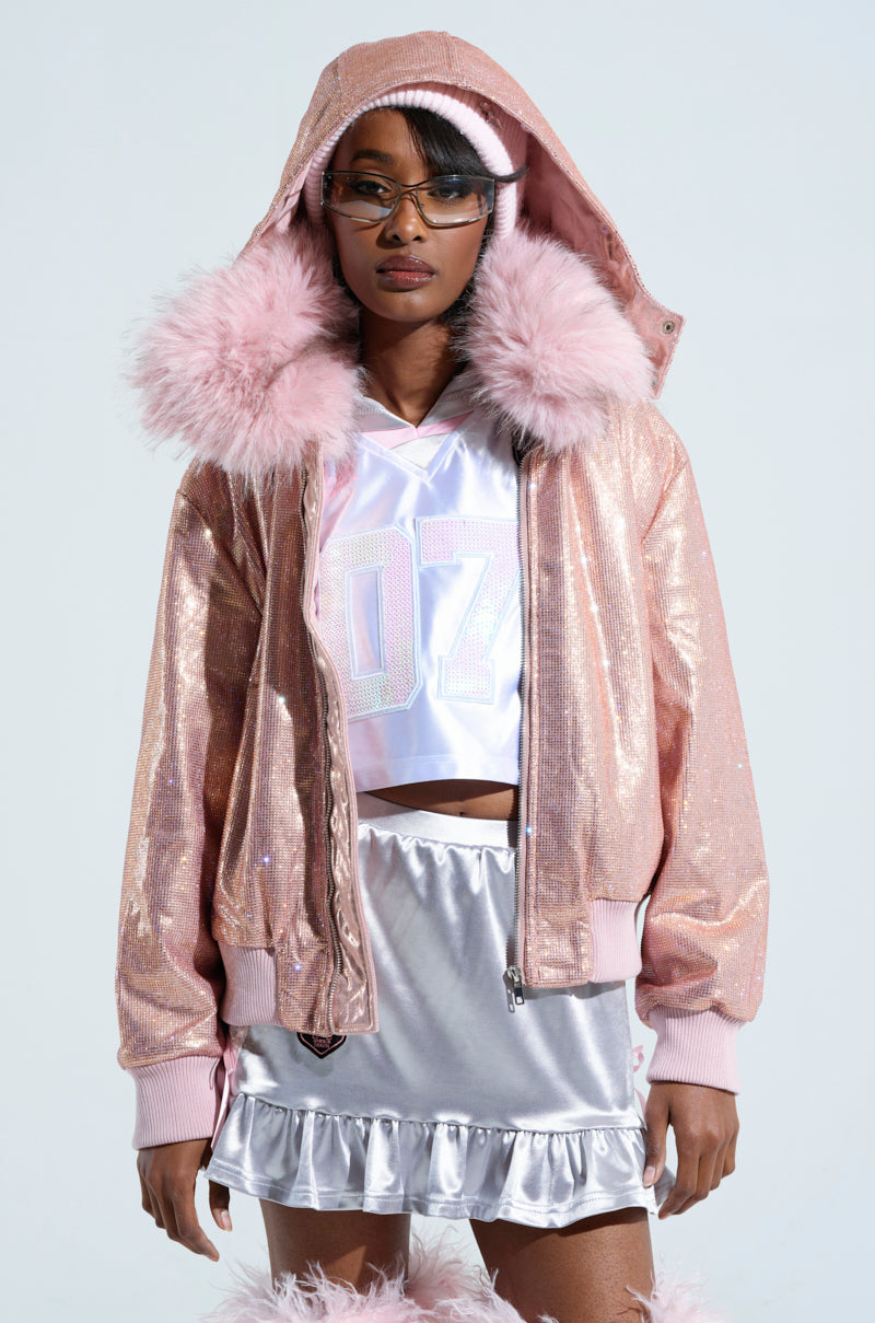 A person wearing the "Eyes on Me Rhinestone Puffer" jacket with a detachable faux fur collar, a white sports jersey, a silver skirt with metallic fabrication, and futuristic sunglasses. The outfit exudes a stylish, futuristic vibe against the simple light gray background.