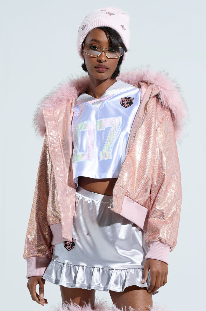 A person wearing the "EYES ON ME RHINESTONE PUFFER" in shiny pink, featuring a detachable faux fur collar, paired with a white jersey displaying "07," a silver ruffled skirt, and accessorized with a pink beanie and glasses. The ensemble merges sporty and glamorous elements for a stylish, futuristic look.